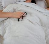 An exhausted woman lays in bed, using one hand to cover her face with a pillow, and the other to hold her glasses on top of the comforter.